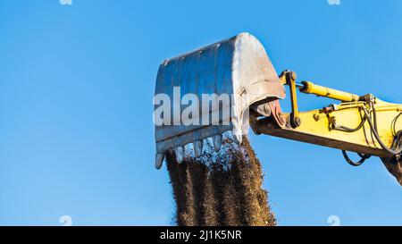 Loose soil falling down of excavator shovel lit by spring sunlight on blue sky background. Closeup of yellow digger arm metal bucket when dumping dirt. Stock Photo