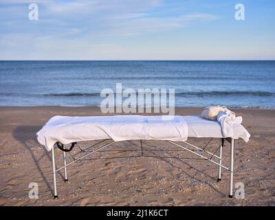 A massage table placed on the sand of a beach with the sea in the background. Stock Photo