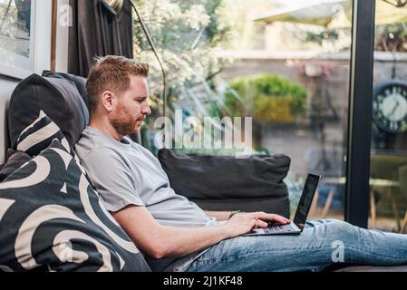 Serious adult man, sitting on couch, using his laptop, browsing online. Stock Photo