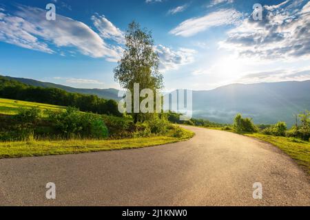 serpentine mountain road down the hill. trees meadow along the way. fog rising in the distant valley. sun above horizon. fluffy clouds on the sky. idy Stock Photo