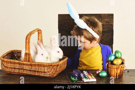 Little child boy in bunny ears with small rabbits in basket and colorful easter eggs sit at table. Stock Photo