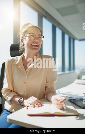 Working with the sun on my back. Shot of a cheerful young businesswoman writing in her notebook while being seated at her desk inside the office Stock Photo