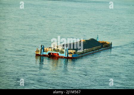 A river non-propelled barge barge (side stone dump vessel) with rubble is anchored in the middle of the river waiting for a tug Stock Photo