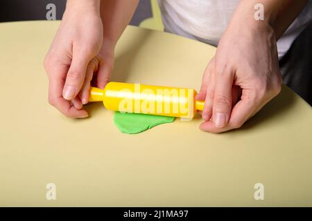 A child and his mother roll out a piece of green dough on the table with a rolling pin, close-up of hands Stock Photo