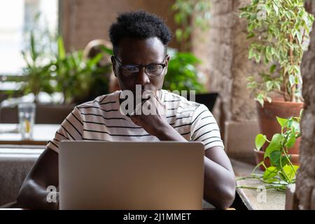 Thoughtful Black man watching educational webinar on laptop, studying, remotely online work in cafe. Stock Photo