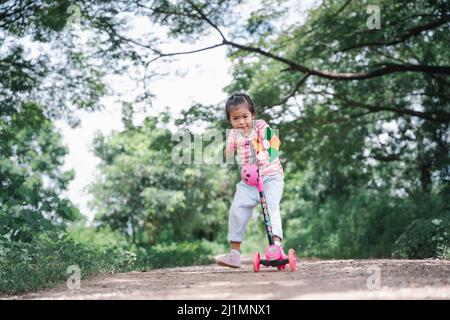 Asia children learn to ride scooters in a park on a summer day. Preschooler girl riding a roller. Kids play outdoors with scooters. Active leisure and Stock Photo