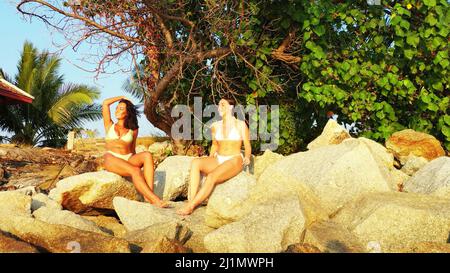 A two Caucasian pretty females in bikinis laying on rocky beach with trees in Koh Samui, Thailand Stock Photo
