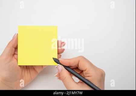 Hand written notes black pencil on yellow sticker. white table background. woman hand writing on yellow sticky notes. female hand holding note paper a Stock Photo