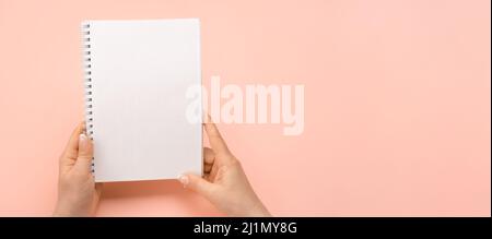 Mockup notepad. Overhead shot of woman hands with blank notepad on pink table. Human hands holding mockup white notebook. Isolated on pink background. Stock Photo