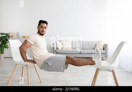 Athletic young Arab man doing strength exercises with two chairs at home Stock Photo