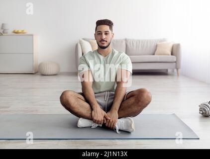 Balanced young Arab guy sitting cross legged on sports mat, meditating or practicing yoga at home, full length Stock Photo