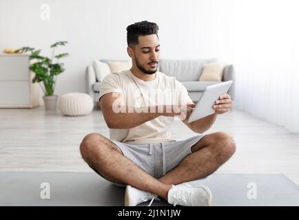 Young Arab guy using tablet computer while sitting cross legged on yoga mat at home Stock Photo