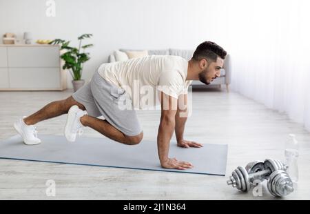 Handsome young Arab guy in sportswear exercising on yoga mat, doing domestic fitness, working out his muscles at home Stock Photo