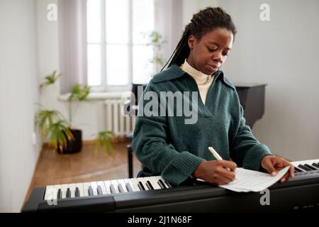 African young woman making notes on paper while sitting at synthesizer, she composing the music at classroom Stock Photo