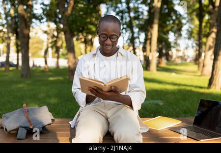 Male african american student reading book outdoors, preparing for classes, sitting in park or university campus Stock Photo