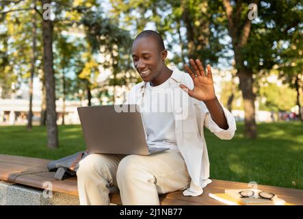 African american male student with laptop attending online lecture, waving hand at webcamera, sitting outdoors Stock Photo