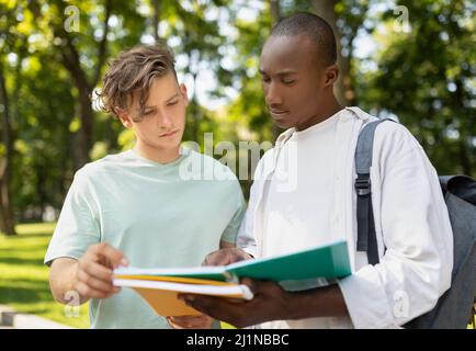 Multiracial university male friends studying together outdoors, standing with workbooks in college campus Stock Photo