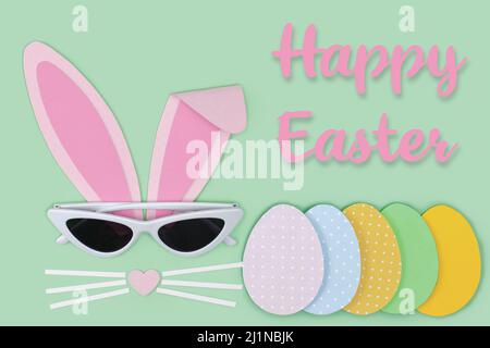Easter greeting card. Bunny rabbit face made of pink paper with sunglasses and painted Easter eggs cutting out paper on blue background. Happy Easter Stock Photo