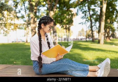 Cute indian female student writing in notebook outdoors, studying online while spending free time in park Stock Photo