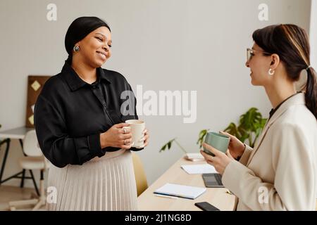 Minimal portrait of two successful young women chatting at coffee break in office, focus on black businesswoman smiling Stock Photo