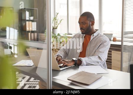 Portrait of adult black businessman using laptop while working in cubicle at modern office Stock Photo