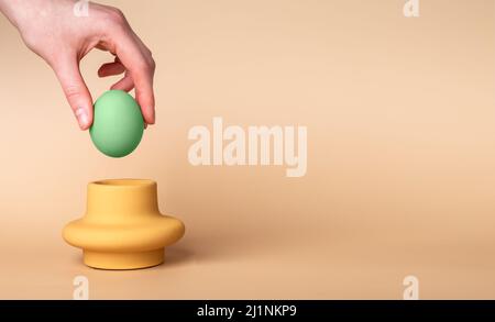 Banner with hand putting green dyed Easter egg in cup. Egg holder and server on beige background. Symbol of Easter holiday. Copyspace. High quality photo Stock Photo