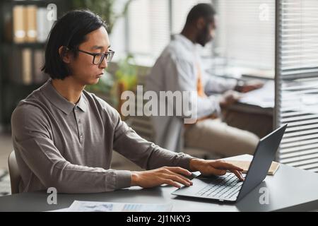 Side view portrait of Asian man in casual wear using laptop while working in office cubicle, copy space Stock Photo