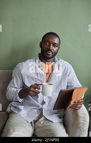 Minimal vertical portrait of handsome African American man holding coffee mug and looking at camera while relaxing on couch against green wall Stock Photo