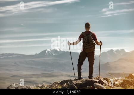 Tourist with backpack and hiking poles stands in mountains and looks at the view Stock Photo