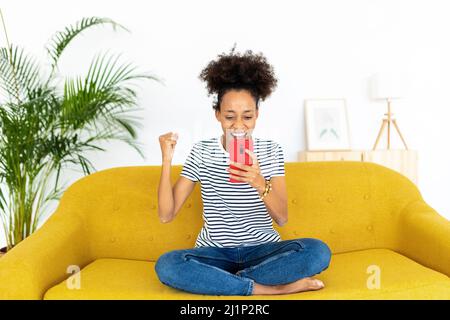 Happy multiracial woman looking at the smartphone and celebrating at the sofa Stock Photo