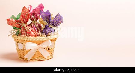 Easter decorative eggs wrapped in color paper with golden ribbons in basket decorated by bow. Pastel pink backdrop. Easter banner. Holiday decorations Stock Photo