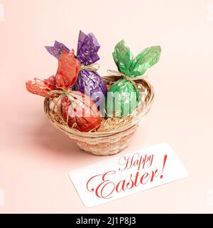 Small basket with three decorative eggs wrapped in color patterned paper and note with words Happy Easter on pastel pink background. Square greeting c Stock Photo