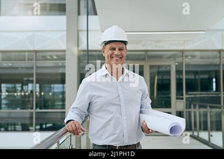 The boss with the plan. Portrait of a cheerful professional male architect looking at the camera while holding blueprints inside a building. Stock Photo