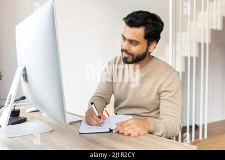 Concentrated positive Indian man thinking, taking notes while sitting at the table with computer at home. Author writing idea for his book, study online. E-learning concept Stock Photo