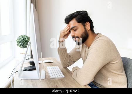 Tired mixed-race hindu male entrepreneur feel eye strain from online work with laptop, indian man sits at the table and massaging eyes, feels headache and lack of energy Stock Photo