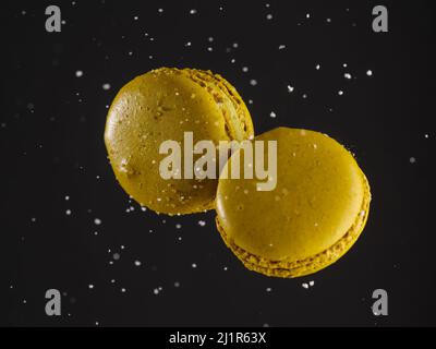 Two sweet yellow macaroons on a black background with grains of sugar in free flight. Festive composition. Birthday, holiday, sweet food, calories. Ba Stock Photo
