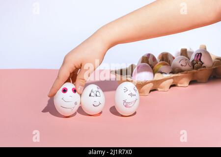 The child puts painted white eggs on a pink background. Painted faces by marker with different happy emotions. Minimal concept, and holiday and needlework. Copy space. Stock Photo