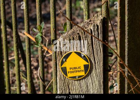Single arrow disc public footpath sign yellow on black pointing straight ahead on a timber post with heavy metal bars in the background Stock Photo