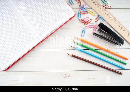From above colored pencils and open notebook arranged on wooden table near paper clips and stapler with ruler for studies Stock Photo
