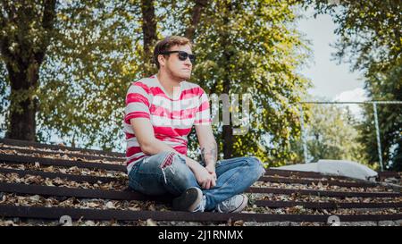 A young handsome man in casual clothes is sitting on the steps in the park and looking around Stock Photo