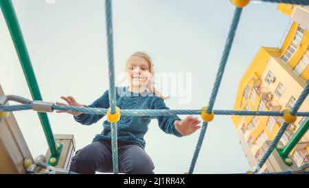 A charming little girl in casual clothes sits on the grid and looks at camera on playground Stock Photo