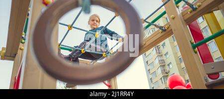 A charming little girl in casual clothes sits on the grid and looks at camera on playground Stock Photo