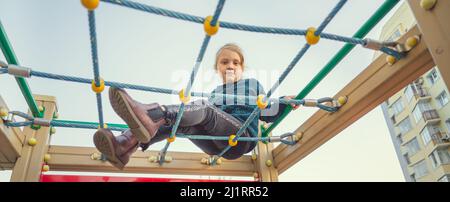 A charming little girl in casual clothes sits on the grid and looks at camera on playground Stock Photo