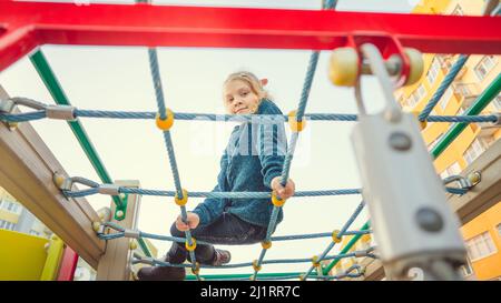 A charming little girl in casual clothes sits on the grid and looks at camera on playground Stock Photo