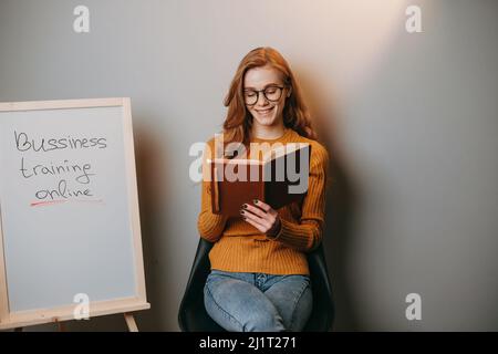 Woman taking online classes on business sitting on chair near a whiteboard and holding a book in her hands. Happy literacy day. Happy teacher day. Stock Photo
