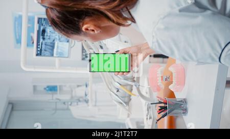 Vertical video: Woman vertically holding mobile phone with green screen in dental cabinet. Dentist looking at isolated mockup background with chroma key on display for dentistry. Stock Photo