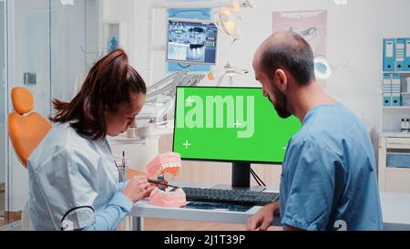 Oral care team analyzing teeth layout while using computer with horizontal green screen on desk. Dentist and dental assistant working with tools and equipment for dentistry and teethcare Stock Photo