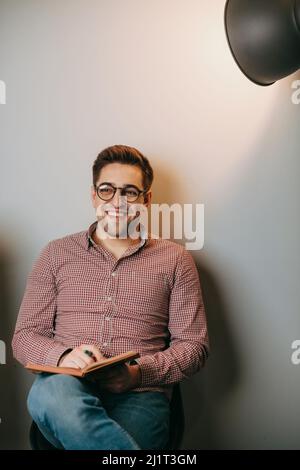 Man reading book while sitting on chair against white wall background. Smiling caucasian teacher wearing eyeglasses. Portrait on light backdrop. Wall Stock Photo