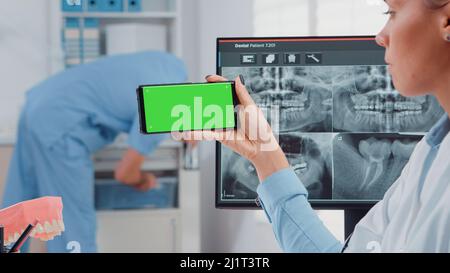 Close up of dentist analyzing smartphone with green screen and teeth scan on computer in dental cabinet. Woman holding device with chroma key for isolated mockup template for dentistry Stock Photo