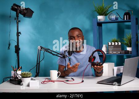 Vlogger sitting at desk in studio with laptop and video recording setup reviewing a pair of audio headphones explaining features. Social media influencer presenting gaming headset to audience. Stock Photo